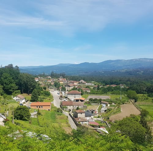 Vista del pueblo de Guláns en los alrededores de la VUT 'O Eido de Celia', ubicado en Ponteareas, Pontevedra. La imagen muestra casas tradicionales rodeadas de campos verdes y caminos rurales, con montañas en el horizonte bajo un cielo despejado. Este pintoresco paisaje gallego refleja la tranquilidad de la vida rural, ideal para quienes buscan desconectar en un entorno natural. Más información en www.oeidodecelia.com.
