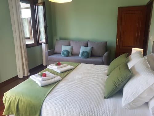 Habitación en la VUT 'O Eido de Celia' en Guláns, Ponteareas, Pontevedra, con una cama doble decorada con ropa de cama en tonos blancos y verdes, acompañada de toallas adornadas con flores frescas. Frente a la cama, un sofá gris con cojines verdes ofrece un espacio adicional para el descanso. Las paredes en tono verde claro y las amplias ventanas llenan la habitación de luz natural, creando un ambiente tranquilo y acogedor. Más información en www.oeidodecelia.com.
