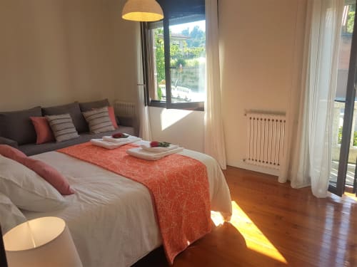 Habitación doble en la VUT 'O Eido de Celia' en Guláns, Ponteareas, Pontevedra. La habitación cuenta con una cama matrimonial decorada con ropa de cama blanca y un cubrecama en tonos naranjas, acompañada de cojines a juego que aportan calidez al ambiente. Un sofá gris con cojines decorativos completa el espacio. Grandes ventanas permiten la entrada de luz natural, creando un ambiente luminoso y acogedor. Más información en www.oeidodecelia.com.