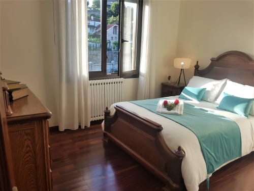 Habitación doble en la VUT 'O Eido de Celia' en Guláns, Ponteareas, Pontevedra. La habitación cuenta con una cama de estilo clásico, vestida con ropa de cama blanca y detalles en turquesa, decorada con un arreglo floral sobre las toallas. La estancia también incluye una cómoda de madera y una lámpara de mesa de luz cálida, creando un ambiente acogedor y sereno. Grandes ventanas permiten la entrada de luz natural, iluminando el espacio y ofreciendo vistas al entorno rural. Más información en www.oeidodecelia.com.