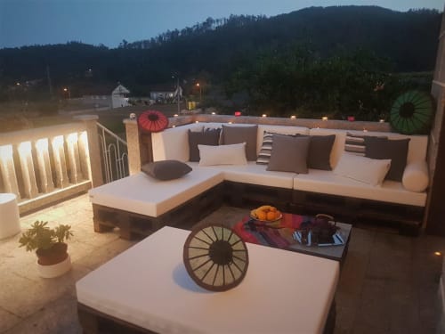 Terraza iluminada al anochecer en la VUT 'O Eido de Celia' en Guláns, Ponteareas, Pontevedra. La terraza está decorada con un sofá en forma de L hecho de palets y cojines en tonos grises y blancos. En la mesa hay una bandeja con frutas frescas y bebidas sobre una manta de colores, acompañada de sombrillas decorativas que aportan un toque original. Las luces cálidas y el entorno natural crean un ambiente acogedor para disfrutar al aire libre. Más información en www.oeidodecelia.com.