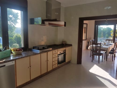 Cocina luminosa en la VUT 'O Eido de Celia' en Guláns, Ponteareas, Pontevedra. La cocina cuenta con una amplia encimera de granito equipada con electrodomésticos modernos y utensilios básicos. A la izquierda, una ventana grande permite la entrada de luz natural y ofrece vistas al exterior. A través de una puerta, se accede a un comedor con una mesa dispuesta para una comida, creando un espacio acogedor y funcional. Más información en www.oeidodecelia.com.
