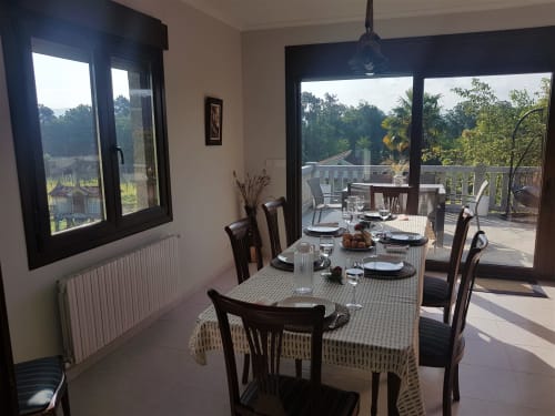 Comedor amplio y luminoso en la VUT 'O Eido de Celia' en Guláns, Ponteareas, Pontevedra. La mesa está dispuesta para una comida, con manteles individuales, platos y copas. Grandes ventanales y una puerta de cristal que da acceso a una terraza permiten la entrada de luz natural y ofrecen vistas al paisaje verde del entorno. Este espacio combina elegancia y conexión con la naturaleza, ideal para disfrutar de comidas en un ambiente relajado. Más información en www.oeidodecelia.com.