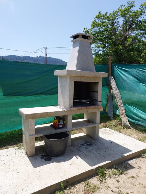 Barbacoa de piedra en el exterior de la VUT 'O Eido de Celia' en Guláns, Ponteareas, Pontevedra. La barbacoa cuenta con estantes laterales para colocar utensilios y una zona de almacenamiento inferior. Está rodeada de un entorno natural, ideal para disfrutar de comidas al aire libre en un ambiente rural y tranquilo. Más información en www.oeidodecelia.com.