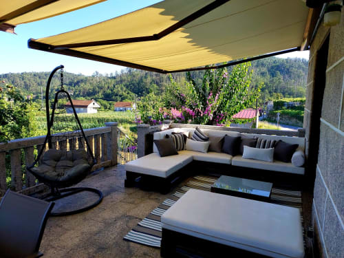 Terraza al aire libre en la VUT 'O Eido de Celia' en Guláns, Ponteareas, Pontevedra, equipada con un cómodo sofá en forma de L, cojines decorativos y una mesa de centro. La terraza también cuenta con una silla colgante y una sombrilla retráctil que proporciona sombra, creando un espacio ideal para relajarse y disfrutar del entorno natural. Al fondo, se aprecian áreas verdes y vistas al paisaje gallego. Más información en www.oeidodecelia.com.