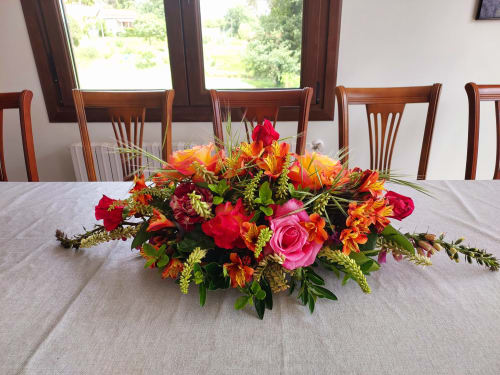 Centro de mesa floral en la VUT 'O Eido de Celia' en Guláns, Ponteareas, Pontevedra. El arreglo incluye una vibrante combinación de flores en tonos rojos, naranjas y rosas, que añade un toque de color y elegancia a la mesa del comedor. Al fondo, las ventanas permiten la entrada de luz natural, destacando el ambiente acogedor del espacio. Más información en www.oeidodecelia.com.