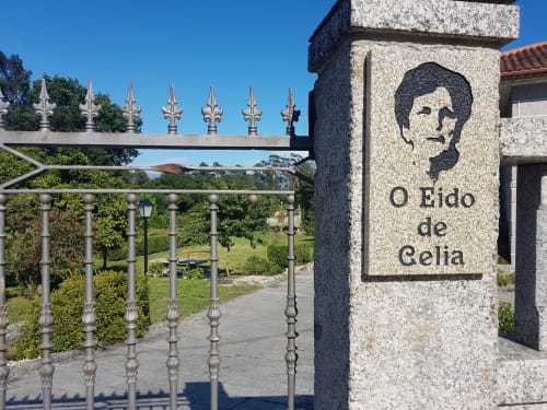 Entrada de la VUT 'O Eido de Celia' en Guláns, Ponteareas, Pontevedra, con una placa de piedra que muestra el nombre del alojamiento y una ilustración en relieve. La puerta de hierro forjado, con detalles decorativos, da paso a los jardines de la propiedad, enmarcando el entorno verde y natural que rodea la casa rural. Más información en www.oeidodecelia.com.