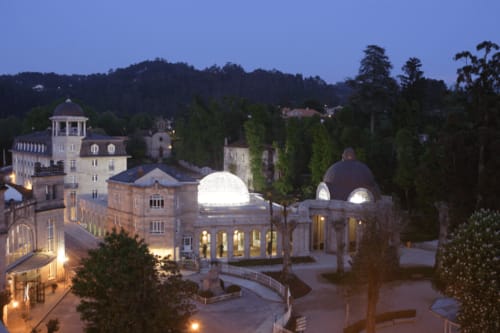 Vista nocturna del balneario de Mondariz en Galicia, con sus icónicas cúpulas iluminadas que destacan en el complejo arquitectónico. Rodeado de frondosa vegetación y colinas, el balneario es uno de los destinos termales más reconocidos de la región. Las luces realzan la elegancia del edificio histórico en contraste con el cielo oscuro, creando un ambiente tranquilo y majestuoso.