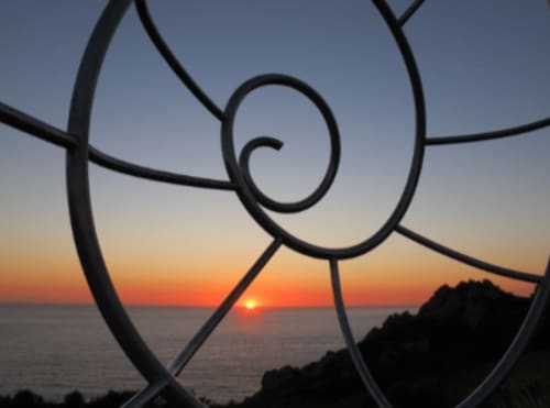 Puesta de sol vista a través de la famosa escultura de la Caracola de Donón en Cangas, Galicia. La espiral de metal enmarca el horizonte mientras el sol desciende sobre el océano, llenando el cielo de tonos cálidos de naranja y rojo. Esta escultura se encuentra en un mirador costero que ofrece vistas impresionantes, combinando arte y naturaleza en un entorno espectacular.