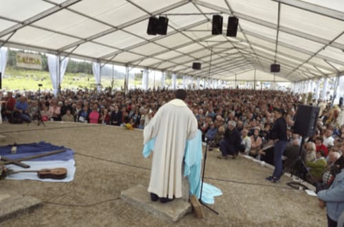 Celebración religiosa en una gran carpa, con un sacerdote de espaldas frente a una multitud de personas asistentes. La estructura cubierta permite reunir a un gran número de participantes para la ceremonia al aire libre. Elementos como guitarras y telas en el escenario sugieren un ambiente de recogimiento y participación comunitaria en el evento.
