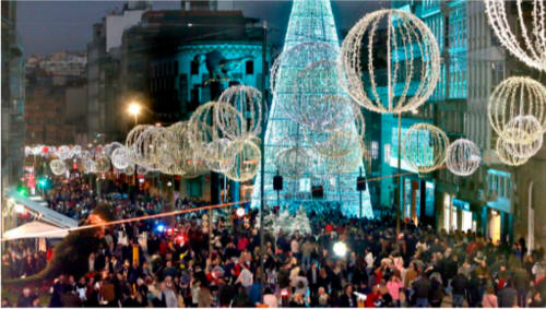 Encendido de luces navideñas en Vigo, Galicia, con una gran multitud de personas llenando las calles decoradas. La imagen muestra un árbol de Navidad iluminado en el centro y grandes esferas de luz suspendidas sobre la avenida, creando un ambiente festivo y animado. La ciudad se ha vuelto famosa por sus elaboradas decoraciones navideñas, atrayendo a numerosos visitantes durante la temporada.