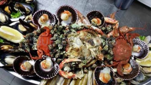 Bandeja de mariscos gallegos variados que incluye navajas, vieiras, percebes, mejillones, camarones y cangrejos, acompañados de rodajas de limón. Esta selección de mariscos frescos es típica de Galicia y destaca por su sabor y calidad, siendo un plato emblemático en la gastronomía de la región.