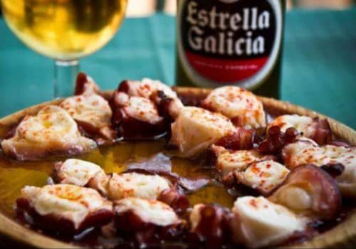 Plato de pulpo a la gallega servido en una tabla de madera, acompañado de pimentón, sal gruesa y un chorrito de aceite de oliva. Al fondo se aprecia una cerveza Estrella Galicia, una combinación clásica para disfrutar de la gastronomía tradicional gallega. Este plato es emblemático de la cocina de Galicia y se sirve comúnmente en festivales y reuniones familiares.