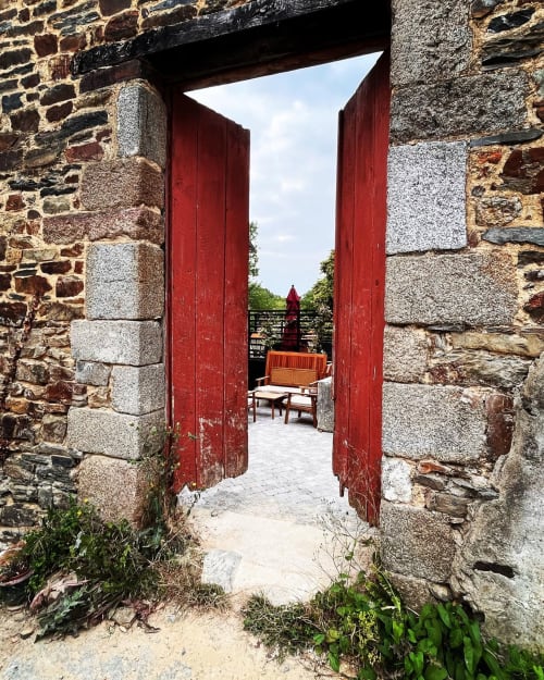 Red wooden doors open to a stone terrace at Domaine du Tertre Ychot, Dol de Bretagne, with garden views beyond