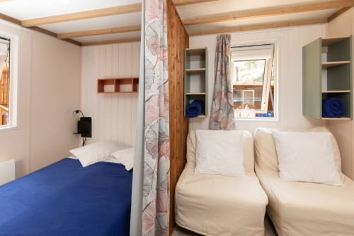 Chambre douillette du Camping La Lézardière avec lits bleu et crème, poutres en bois, étagères et fenêtre sur jardin