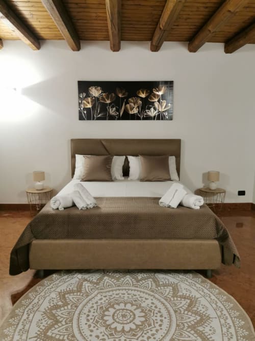 Elegant bedroom at Casa Bellini Palermo with taupe upholstered bed, white pillows, wooden beams, mandala rug, and floral