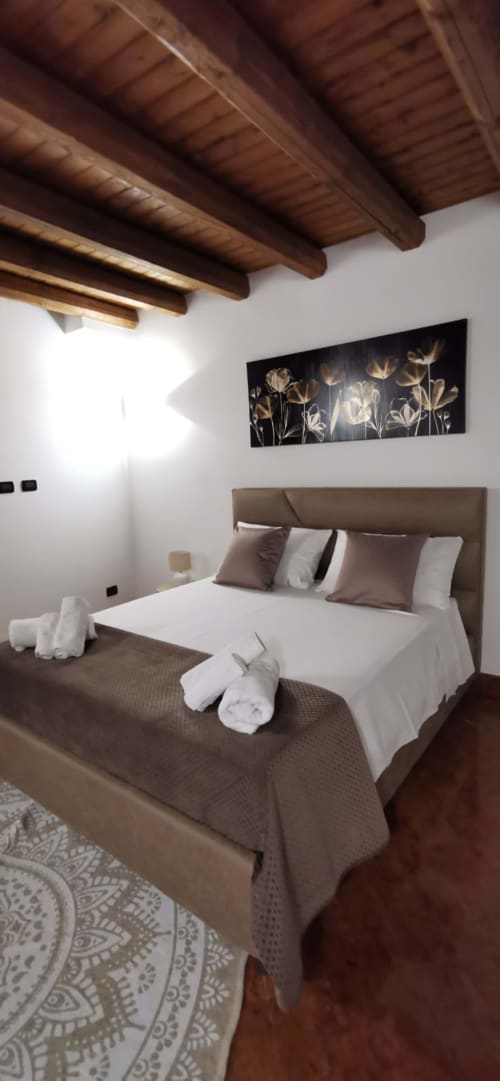 Spacious bedroom at Casa Bellini Palermo with exposed wooden beams, taupe headboard, white linens, and floral wall art