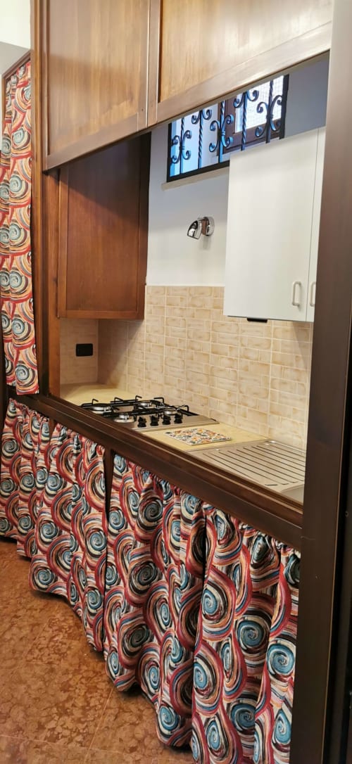 Compact kitchenette at Casa Bellini in Palermo with gas stove, beige tile backsplash, wooden cabinets, and patterned curtain