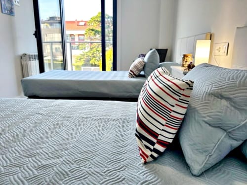 Moderno dormitorio en apartamento de Baiona con dos camas, almohada a rayas y amplios ventanales con vistas a edificios de la ciudad