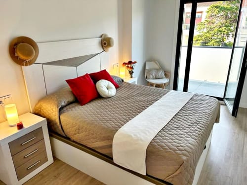 Dormitorio moderno en apartamento de Baiona con cama, almohadas rojas, mesita de noche, lámpara y acceso a balcón con vistas a la piscina
