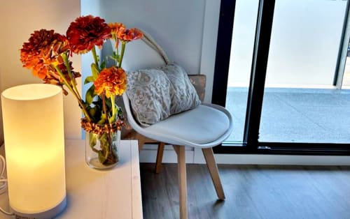 Rincón moderno de apartamento en Baiona con gerberas rojas, lámpara cilíndrica, silla y vistas al océano a través de puertas de cristal