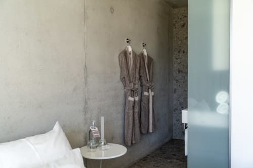 Minimalistische Schlafzimmer im Le Pavillon M in Grignan mit Betonwänden, weißen Kissen und grauen Bademänteln an Haken