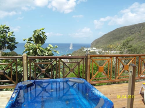 Piscine bleue sur terrasse en bois surplombant la baie de Deshaies avec voilier, collines verdoyantes et végétation tropicale