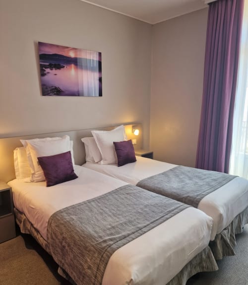 Due letti singoli con biancheria bianca e accenti viola in una camera moderna dell'Hôtel De La Fontaine a Nizza, Francia
