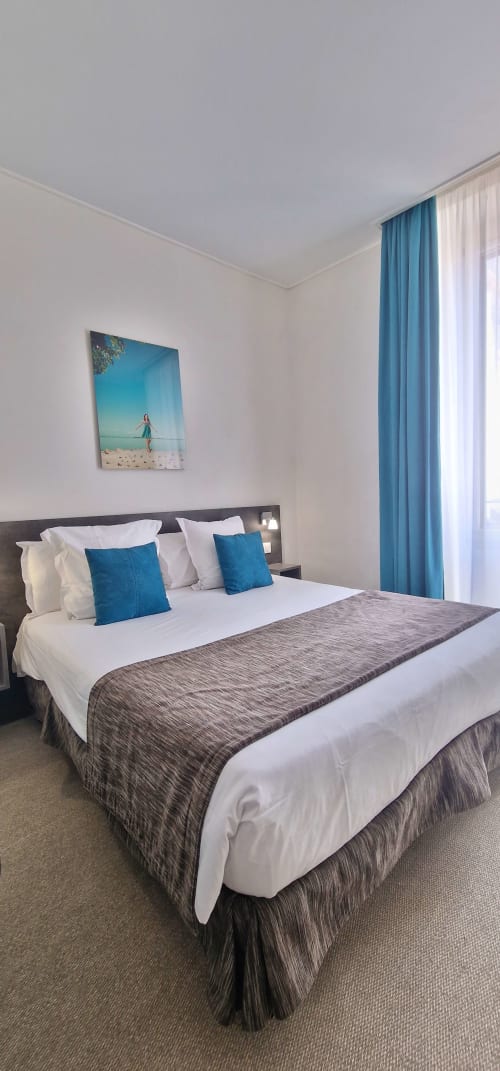 Modernes Hotelzimmer mit blauen Kissen, weißen Betttüchern, Strandkunst und türkisen Vorhängen im Hôtel De La Fontaine in Nizza