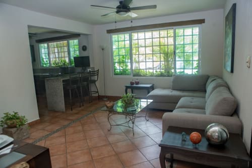 Amplia sala de villa con sofá seccional gris, grandes ventanales con vistas al jardín tropical, ventilador de techo y planta abierta