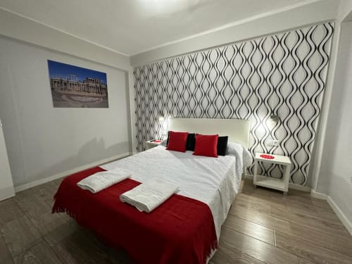 Moderna habitación del apartamento Gestyp-AT en Mérida con cama blanca, camino rojo, cabecero con papel tapiz estampado y vista a la ciudad