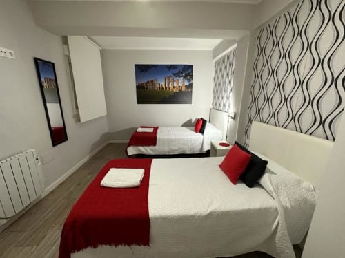 Dormitorio doble en apartamento Gestyp-AT en Mérida con camas blancas, acentos rojos, papel tapiz a rayas y cuadros de vistas de la ciudad.