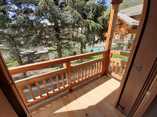 Sunny wooden balcony with mountain views at Serre Chevalier Cristal Lodge, featuring terrace seating and forest surroundings.