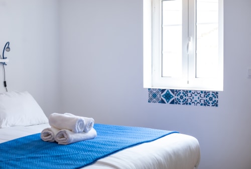 Quarto Santo António na pousada Ó da Casa em Peniche com roupa de cama azul, toalhas enroladas e janela com azulejos padronizados azuis