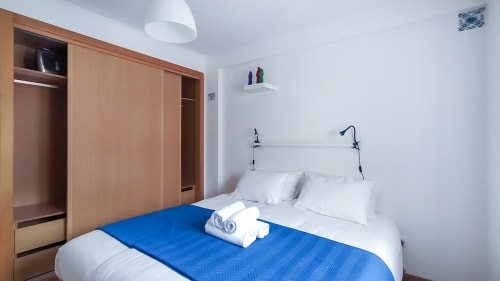 Quarto iluminado em Ó da Casa em Peniche com manta azul, roupa de cama branca, guarda-roupa em madeira e prateleiras de parede