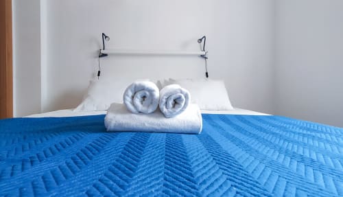 Cama impecavelmente arrumada com colcha acolchoada azul, almofadas brancas, toalhas enroladas e cabeceira metálica no Ó da Casa B&B em Peniche
