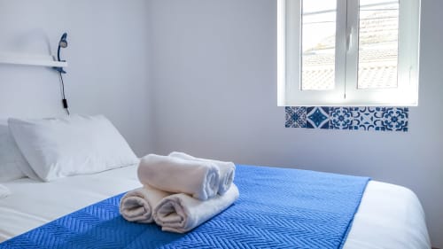 Quarto limpo na pousada Ó da Casa em Peniche com colcha azul, toalhas enroladas, travesseiro branco e janela com vista para a cidade