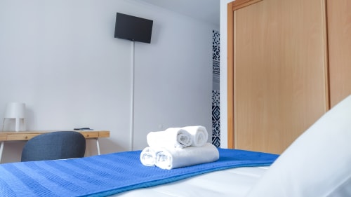 Quarto moderno em Ó da Casa em Peniche com roupa de cama azul, toalhas brancas enroladas, TV na parede, mesa e cadeira de madeira