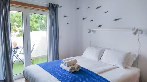 Quarto ensolarado na Ó da Casa em Peniche com toque azul, roupa de cama branca, acesso à varanda e arte com peixes.