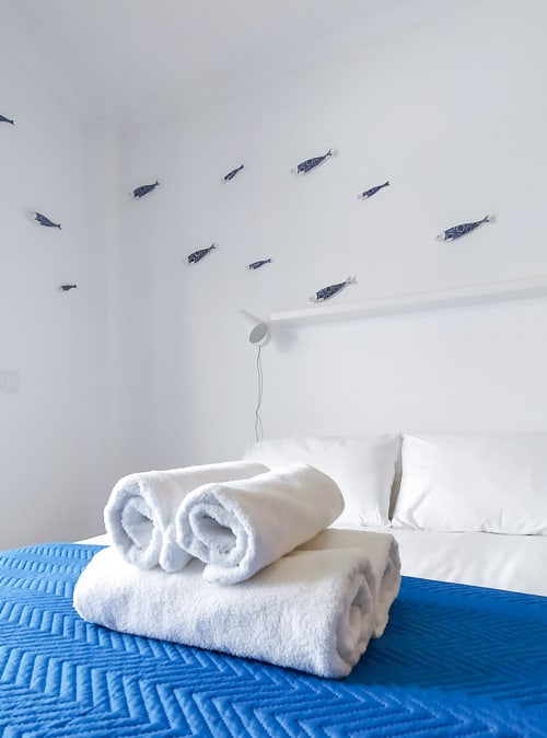 Toalhas brancas enroladas sobre cama azul no quarto Sardinha da Ó da Casa, pousada em Peniche com decoração de peixes na parede
