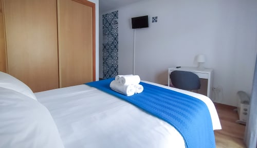 Quarto Sardinha em Ó da Casa em Peniche com cama branca, manta azul, toalhas enroladas, televisão de tela plana e guarda-roupa de madeira