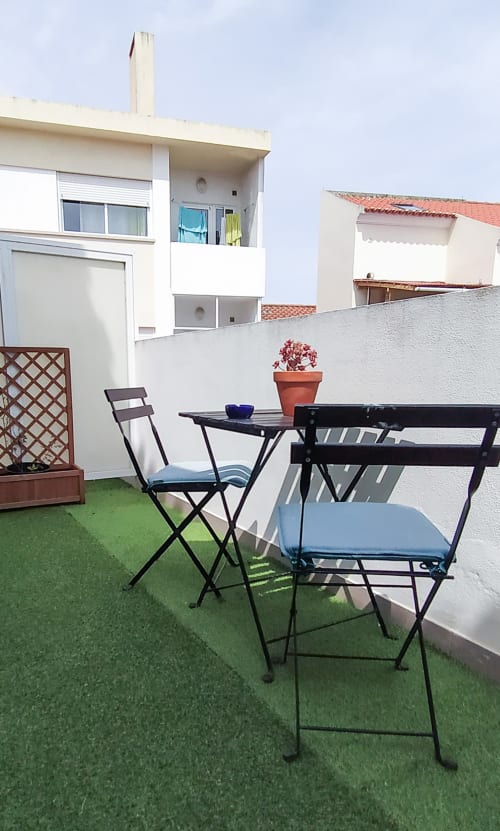 Aconchegante terraço da pousada Ó da Casa em Peniche com cadeiras dobráveis pretas, mesa e vasos de flores com vista para a cidade