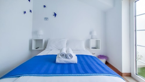Quarto luminoso em Ó da Casa em Peniche com edredom azul, travesseiros brancos, toalhas enroladas e decorações de pássaros azuis