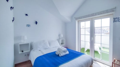 Quarto luminoso na Ó da Casa em Peniche com colcha azul, roupa de cama branca, portas de vidro para a varanda e parede com pássaro azul