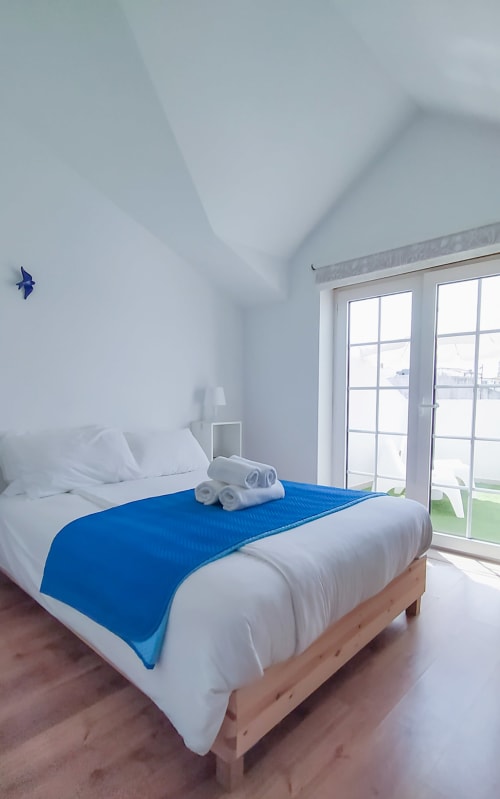 Quarto moderno em Ó da Casa em Peniche com caminho azul sobre roupa de cama branca, estrutura em madeira e portas-janelas de vidro com vista