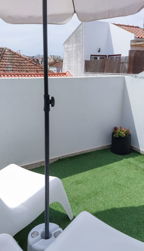 Varanda de cobertura Peniche BnB com grama artificial, guarda-sol, cadeiras brancas e vista para telhados de terracota
