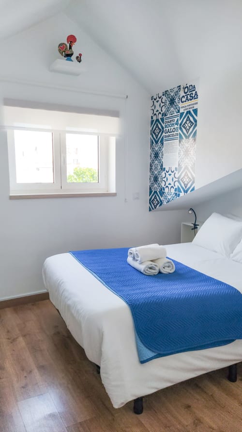 Quarto brilhante em Ó da Casa em Peniche com cama branca, manta azul, vista pela janela e pôster de azulejos portugueses