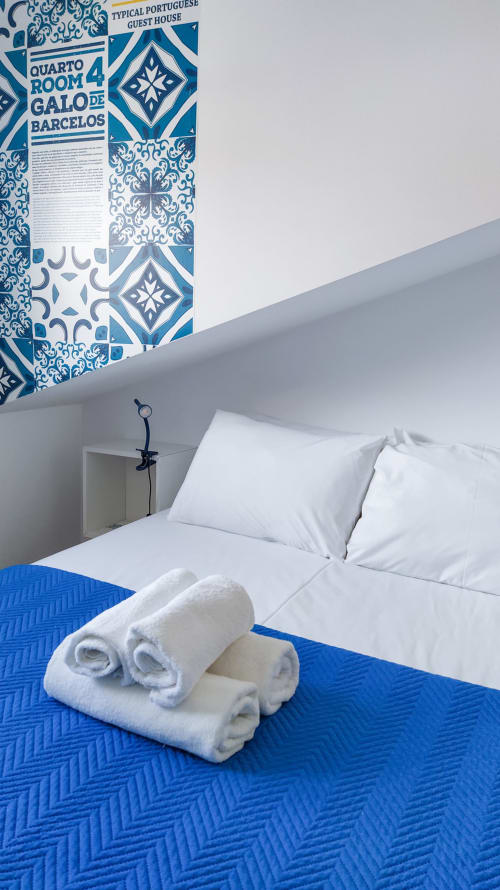 Quarto Galo de Barcelos na pousada Ó da Casa em Peniche com cama branca, passadeira azul, toalhas enroladas e parede de azulejos portugueses
