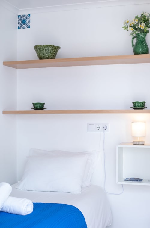 Quarto luminoso em Ó da Casa em Peniche com prateleiras de madeira, plantas em vasos, almofadas brancas e detalhe de manta azul