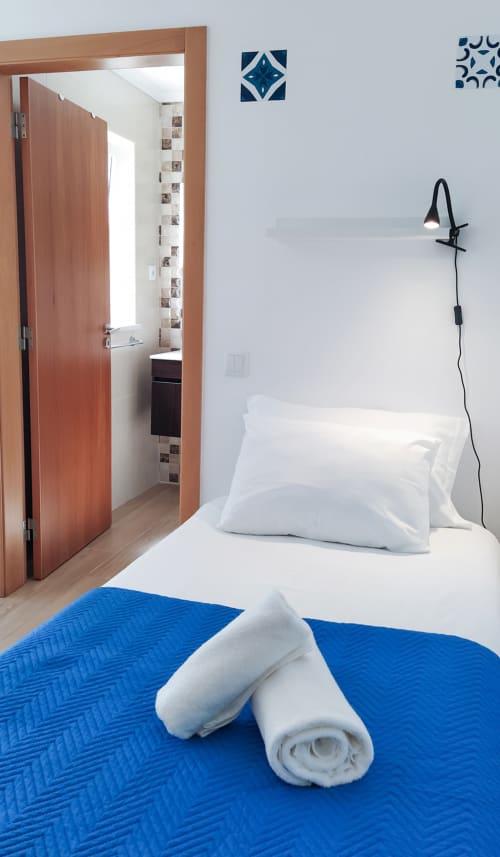 Quarto moderno do hotel Ó da Casa em Peniche com colcha azul, almofadas brancas, toalhas enroladas e casa de banho privativa